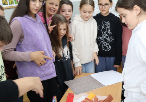 Uczniowie klasy 4 stoją wokół stołu z przyborami plastycznymi i kolorowymi kartkami, rozmawiają i przygotowują materiały do wspólnej pracy w sali lekcyjnej.