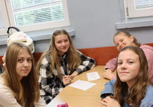 Cztery uczennice klasy 6 siedzą przy stoliku w sali lekcyjnej z kartkami i długopisem, uśmiechają się do aparatu. Na stole stoi różowa butelka na wodę.