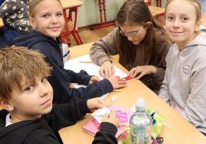 Uczniowie klasy 5 siedzą przy stole i pracują nad zadaniem. Troje z nich uśmiecha się do aparatu, a jedno skupia się na rysowaniu. Na stole leżą przybory i butelka z wodą.