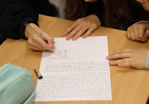 Dłonie uczniów piszących list do patronki szkoły – Stefanii Sempołowskiej. Na stole leży kartka z odręcznym tekstem, błękitny piórnik i skuwka od długopisu.