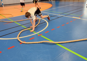 Uczniowie podczas zajęć sportowych w sali gimnastycznej. Chłopiec w białej koszulce ćwiczy z grubą liną na niebieskiej podłodze, w tle kosz do koszykówki i drabinki.