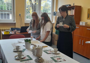 Trzy osoby w sali nauczycielskiej — dwie uczennice przeglądają kartki przy biurku, a nauczycielka z filiżanką stoi obok stołu nakrytego białym obrusem i porcelanową zastawą.