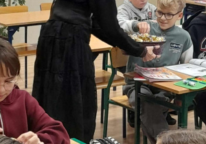Uczennica w czarnej sukience rozdaje cukierki uczniom podczas lekcji języka angielskiego. Dzieci siedzą przy stolikach z otwartymi podręcznikami i piórnikami.