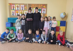 Uczniowie klasy I pozują do zdjęcia z uczennicami klasy 8 – Magdą i Hanią. W tle gazetka z napisem „Jesień” i rysunkami drzew. Jedna z uczennic trzyma kosz z cukierkami.