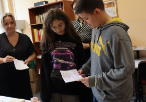 Pani Katarzyna Głuszkiewicz oraz uczniowie klasy 7b, Paulina i Emil, stoją przy stole z książkami i porcelanową zastawą. Uczniowie czytają tekst z kartek w sali szkolnej.