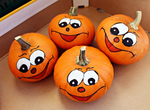 Cztery małe, pomarańczowe dynie z namalowanymi uśmiechniętymi twarzami – duże oczy, czerwone noski i szerokie uśmiechy. Leżą razem w kartonowym pudełku, tworząc wesołą kompozycję.