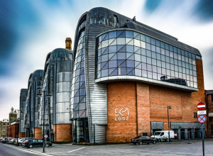 Nowoczesny budynek EC1 w Łodzi z charakterystyczną szklaną i metalową elewacją połączoną z ceglanymi fragmentami dawnej elektrowni. Na ścianie widoczne białe logo „EC1 Łódź”.