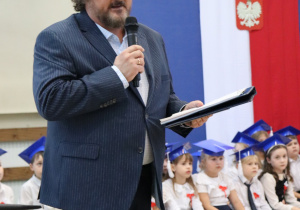 Przedstawiciel Rady Rodziców. W tle widać uczniów w niebieskich biretach oraz biało-czerwoną dekorację z godłem Polski.