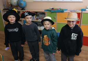 Czworo uczniów klasy 1 stoi w sali lekcyjnej podczas „Dnia kapelusza”. Każde dziecko ma na głowie inne nakrycie — piracki kapelusz, elegancki kapelusz słomkowy, szyty kapelusz z ozdobą. W tle widać mapę Polski oraz szkolne szafki.