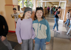 Dwie uczennice stoją na szkolnym korytarzu podczas „Dnia kapelusza”. Jedna ma na głowie szarą czapkę zimową, druga czarny kapelusz typu bucket i okulary. W tle widać innych uczniów uczestniczących w wydarzeniu.