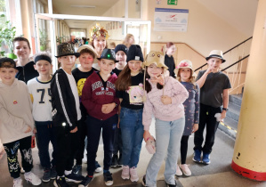 Grupa uczniów stoi na szkolnym korytarzu w ramach „Dnia kapelusza”. Dzieci mają na głowach fantazyjne nakrycia — m.in. kapelusze słomkowe, czapki zimowe, kapelusz z uszami królika oraz kapelusz ozdobiony liśćmi. W tle widać schody i innych uczniów.