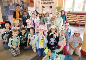 Grupa uczniów klasy 2 pozuje na szkolnym korytarzu wraz z wychowawczynią. Dzieci mają różnorodne kapelusze i czapki – słomkowe, ozdobne, kolorowe – w ramach „Dnia kapelusza”. W tle widać ścianę z dekoracją i tablicę z nazwą szkoły.