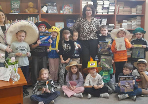 Grupa uczniów klasy 1 pozuje w bibliotece szkolnej z nauczycielem bibliotekarzem Moniką Juran. Dzieci mają na głowach różnorodne kapelusze i trzymają w rękach książki lub zakładki, uczestnicząc w „Dniu kapelusza” połączonym z pasowaniem na czytelników. W tle widać regały z książkami.