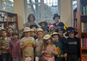 Grupa uczniów klasy 1 pozuje w bibliotece szkolnej razem z nauczycielem bibliotekarzem Moniką Juran i wychowawczynią Anną Wlazeł. Dzieci mają na głowach różnorodne kapelusze — eleganckie, fantazyjne i zabawne — uczestnicząc w „Dniu kapelusza” połączonym z pasowaniem na czytelników. W tle widać regały pełne książek.