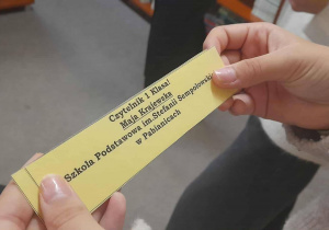 Zbliżenie dłoni dziecka trzymającego imienną żółtą zakładkę do książki z napisem „Czytelnik I Klasa! Maja Krajewska. Szkoła Podstawowa im. Stefanii Sempołowskiej w Pabianicach”. W tle widoczne rozmyte sylwetki innych uczniów.