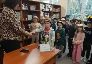 Nauczyciel bibliotekarz stoi przy biurku w bibliotece szkolnej i przyjmuje przysięgę od pierwszoklasisty, który trzyma rękę na książce. Za nim stoi grupa dzieci w kapeluszach uczestnicząca w pasowaniu na czytelników.