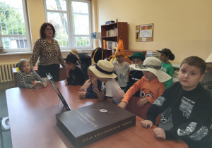 Uczniowie klasy 1 siedzą przy stole w bibliotece szkolnej, słuchając nauczyciela bibliotekarza podczas pasowania na czytelników. Dzieci mają na głowach różnorodne kapelusze, a na stole leży duży „Atlas Rzeczypospolitej Polskiej”.