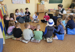 Grupa uczniów siedzi na podłodze w kręgu, słuchając pani z grupy „Miauczykotek”, która trzyma książkę o kotach i prowadzi zajęcia na temat opieki i żywienia kotów. W tle widać szkolną klasę.