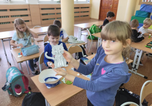 Dziewczynka stoi przy ławce w klasie i trzyma w ręku wycięty papierowy rysunek kota. W tle inni uczniowie pracują nad swoimi pracami plastycznymi związanymi z tematyką kotów.
