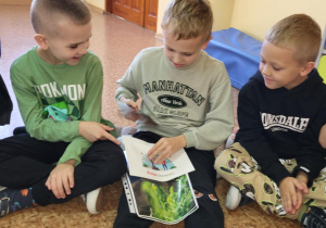 Trzech chłopców siedzi na podłodze w klasie i przegląda kolorowe materiały szkoleniowe o kotach, rozmawiając i wskazując ilustracje.