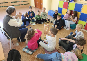 Grupa uczniów siedzi w kręgu na podłodze wokół prowadzącej zajęcia, która pokazuje gestami coś związanego z kotami. Dzieci słuchają uważnie i aktywnie uczestniczą w rozmowie o opiece nad kotami.