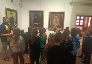 Grupa uczniów stojących przed trzema portretami w muzeum, słuchających przewodnika opowiadającego o obrazach.