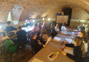 Uczniowie siedzący przy stołach podczas warsztatów plastycznych w muzeum; prowadzący pokazuje duży rysunek postaci.