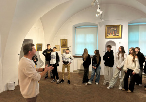 Przewodnik opowiada uczniom o obrazach na wystawie Kislinga w jasnej sali muzealnej; młodzież słucha w skupieniu.