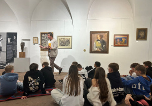 Przewodnik opowiada uczniom siedzącym na podłodze o obrazach i rzeźbach na wystawie Kislinga w Pabianicach.