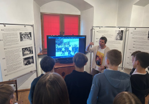 Przewodnik pokazuje uczniom film o pojedynku Kislinga na ekranie podczas lekcji muzealnej, młodzież słucha w skupieniu.