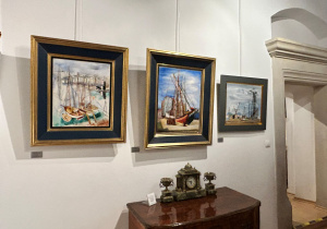 Trzy obrazy Kislinga z motywem statków i portu zawieszone nad zabytkową komodą z zegarem na wystawie w muzeum.
