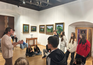 Przewodnik rozmawia z uczniami na tle pejzażowych obrazów Kislinga; młodzież słucha w skupieniu w sali muzeum.