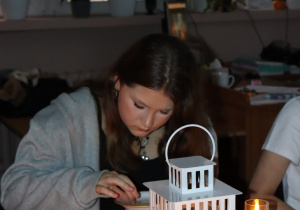 Uczennica klasy 6 siedzi przy świecach i lampionie podczas czytania „Dziadów”, wykonując symboliczny obrzęd z talerzykiem.