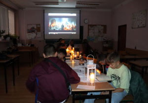 Uczniowie czytają „Dziady” przy świecach, w sali z wyświetloną sceną obrzędową na ekranie, tworząc teatralny klimat wydarzenia.