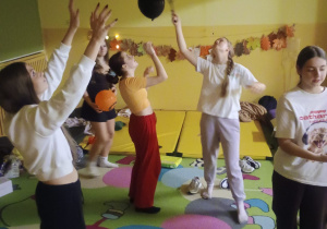 Grupa uczniów klasy 8b stoi w sali szkolnej udekorowanej jesiennymi liśćmi i bawi się balonami w halloweenowych kolorach, odbijając je w górę. W tle widać materace i jesienne dekoracje.