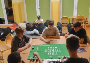 Grupa uczniów klasy 8b siedzi przy stole w szkolnej świetlicy i gra w grę z użyciem żetonów na zielonej macie. W tle widać kilku uczniów siedzących i leżących na materacach przy ścianie.