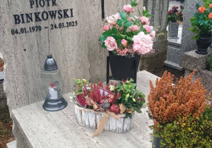 Nagrobek ozdobiony zapalonym zniczem oraz bukietami kwiatów i wrzosów. W tle widoczne są inne pomniki i jesienne drzewa na cmentarzu.