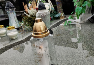 Zapalony znicz stojący na płycie nagrobnej, obok karteczki z napisem „Pamiętamy” od Szkoły Podstawowej nr 13 w Pabianicach; w tle widać dekorowane nagrobki i jesienne drzewa.