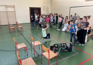 Dzieci na sali gimnastycznej uczestniczą w torze przeszkód, przechodząc pod krzesłami, podczas gdy reszta grupy świetlicowej kibicuje.