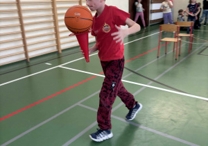 Dziecko biegnie po sali gimnastycznej, trzymając piłkę na pachołku podczas zawodów sportowych w świetlicy.