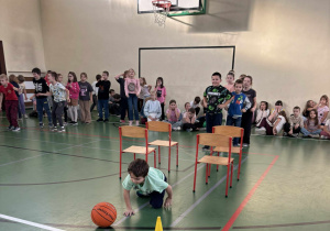 Dziecko czołga się po podłodze sali gimnastycznej, omijając pachołek i piłkę podczas zawodów sportowych, w tle grupa uczniów kibicuje.