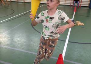 Dziecko na sali gimnastycznej niesie plastikowy pachołek z piłką do koszykówki, wykonując zadanie podczas zawodów sportowych w świetlicy.