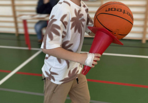 Dziecko na sali gimnastycznej niesie piłkę na plastikowym pachołku podczas konkurencji sportowej w świetlicy.