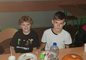 Dwóch uczniów siedzi przy stole podczas halloweenowej zabawy; na stole znajdują się przekąski, napoje i świecąca dynia, a w tle wiszą szkolne plakaty biologiczne.