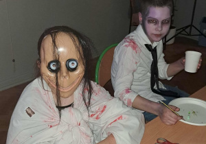 Dwóch uczniów w halloweenowych przebraniach siedzi przy stole; jeden ma straszną maskę z dużymi oczami, a drugi makijaż zombie i poplamioną koszulę. Na stole stoją talerzyki, kubki i butelka soku.
