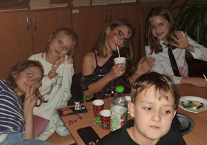 Grupa uczniów w halloweenowych przebraniach siedzi przy stoliku, uśmiecha się i pozuje do zdjęcia; na stole leżą słodycze, kubki i butelka soku. Za nimi widać szafki z modelami biologicznymi.