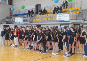 Grupa uczniów w czarnych koszulkach ustawiona na parkiecie hali sportowej podczas charytatywnego meczu wolontariuszy szkół pabianickich, w tle trybuny i banery sponsorów.