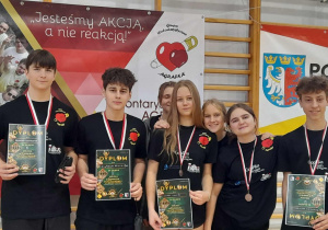 Uczniowie SP 13 w Pabianicach stojący z medalami i dyplomami na tle banerów charytatywnego turnieju w hali sportowej.