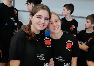 Uczennice SP 13 w Pabianicach uśmiechające się podczas charytatywnego wydarzenia sportowego, w tle pozostali uczniowie w koszulkach wolontariatu.