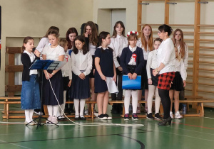 Uczennice w galowych strojach prezentują część artystyczną apelu niepodległościowego na sali gimnastycznej, czytając tekst do mikrofonu pod opieką nauczycielki.